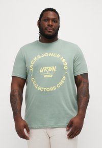 T-shirt in cotone verde con una grafica rotonda in testo giallo che recita "JACK&JONES 1990 URBN COLLECTORS CREW", maniche corte e scollatura rotonda.