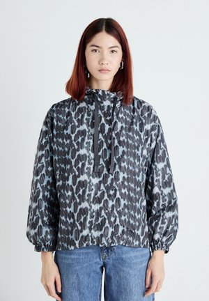 Jeune femme aux cheveux raides et rouge portant une veste à imprimé léopard noir et gris et un jean bleu, se tenant devant un fond blanc uni.