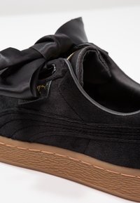 Zblízka pohled na černou semišovou tenisku s velkou saténovou mašlí a hnědou texturovanou podrážkou na bílém pozadí.