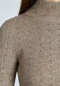 Maglione beige a coste con collo alto, caratterizzato da decorazioni metalliche sparse per aggiungere texture e dettagli.