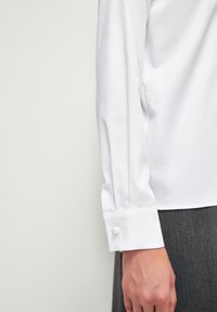 Camicia bianca a maniche lunghe con una texture liscia, caratterizzata da dettagli con polsini bottonati e vestibilità su misura, abbinata a un capo grigio sottostante.