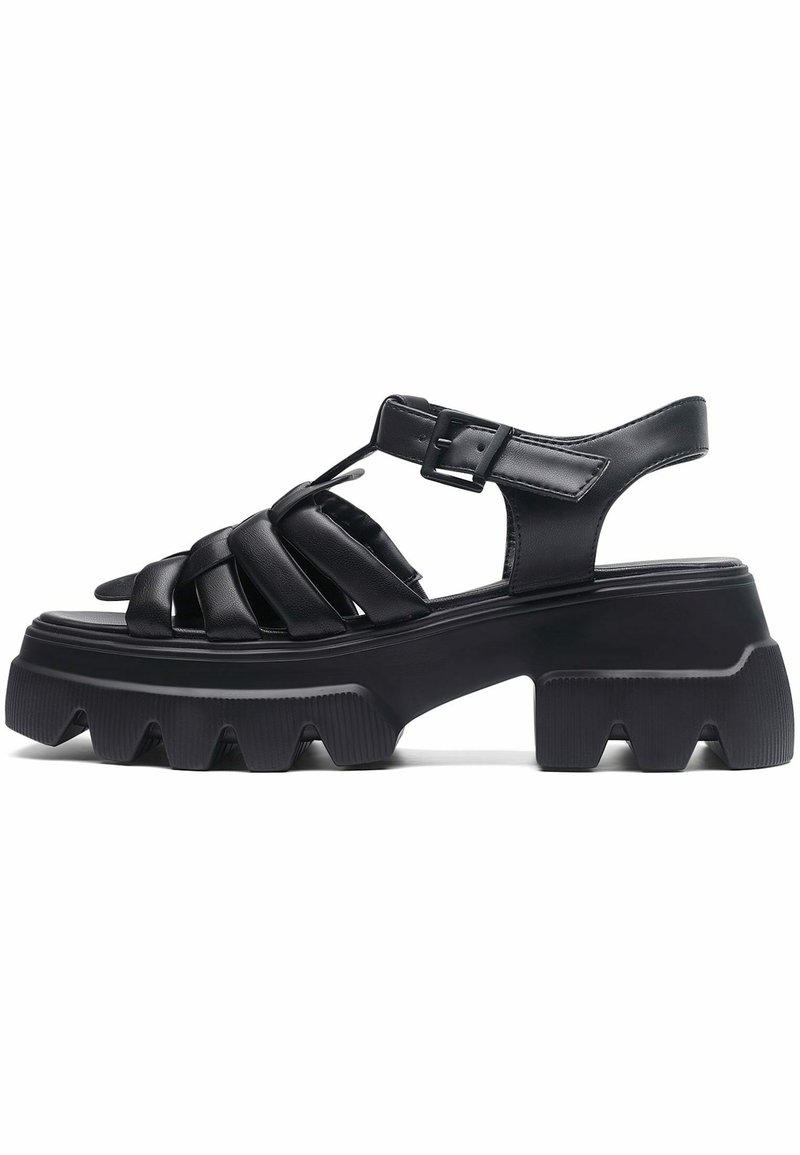 Sandales en cuir noir avec semelles épaisses et rainurées, dotées de lanières entrecroisées et d'une boucle réglable pour un ajustement sécurisé. Dessus ouvert à l'arrière.