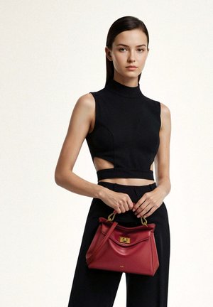 Jeune femme portant un haut sans manches noir et un pantalon, tenant un sac à main en cuir rouge avec des accessoires dorés devant un fond uni.