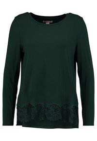 Langärmliges, dunkelgrünes Oberteil aus weichem Material, mit einem gewellten Saum und aufwendigen schwarzen Spitzenverzierungen am unteren Rand.