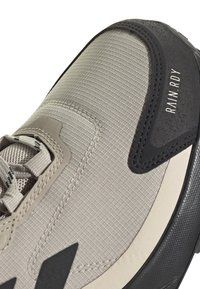 Bottine de sport gris clair avec tissu texturé, accents en caoutchouc noir et semelle foncée contrastante. Présente des détails de couture et un logo.