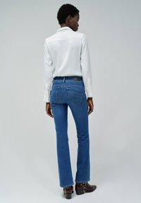 Jeans ajustados de color azul con un ligero acampanado, cintura alta y dos bolsillos traseros. Combinados con una camisa blanca de cuello y botas de tacón marrones.