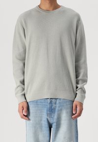 Man in een lichtgrijze, gebreide trui met lange mouwen en vervaagde blauwe distressed jeans, staand tegen een effen achtergrond.