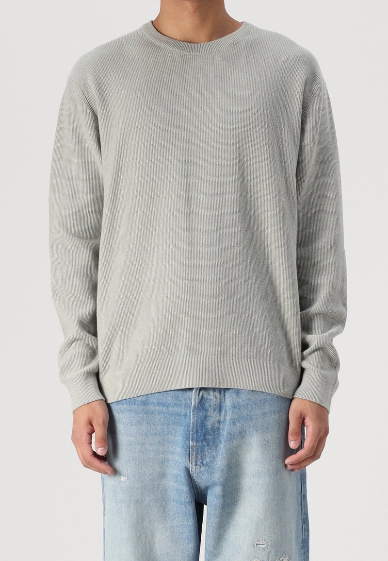 Man in een lichtgrijze, gebreide trui met lange mouwen en vervaagde blauwe distressed jeans, staand tegen een effen achtergrond.