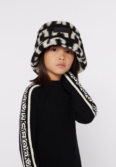 Zwart-wit gestreepte bucket hat van zacht nepbont, met een logo-opdruk op de voorkant, gedragen met een zwart sweatshirt met witte strepen.