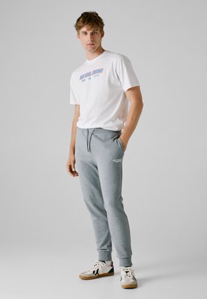 Witte t-shirt met korte mouwen met afdruk "Red Bull Racing"; grijze joggingbroek met zijzakken; beige en zwarte sneakers.