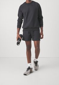 Homme portant un sweat-shirt noir et un short, tenant une bouteille d'eau noire, portant des chaussettes blanches Nike et des chaussures de sport grises.