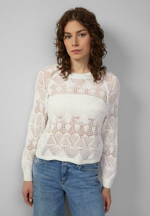 Weißer Strickpullover mit einem spitzenähnlichen Muster, mit einem runden Ausschnitt und gerippten Bündchen. Kombiniert mit hellblauen Jeans.