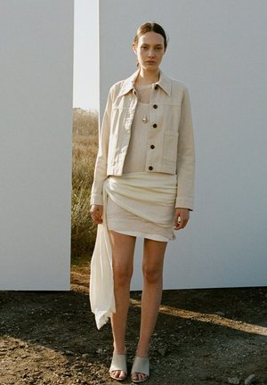 Mujer de pie al aire libre entre paneles blancos, vistiendo una chaqueta beige, vestido claro con textura y tela drapeada, collar con colgante y mules abiertos en los dedos.