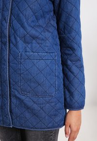 Blå quiltad jacka i denimtyg, med framsidig dragkedja och en enda ficka med diamantmönster i sömmen.