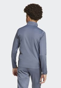 Chaqueta atlética gris con un cuello alto y mangas largas, con una textura suave. Combinada con pantalones a juego, mostrada desde la parte trasera.