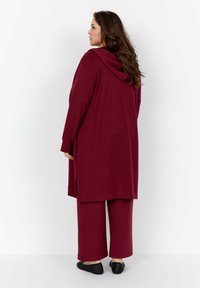 Eine Frau steht, trägt eine lange burgunderfarbene Kapuzenjacke und passende weit geschnittene Hosen mit schwarzen Slippers und steht seitlich.