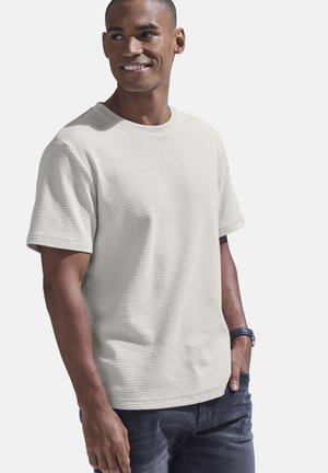 Light beige short-sleeve textured t-shirt with a crew neckline, worn with dark jeans, featuring a casual fit and visible stitching details.