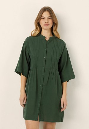 Abito verde a maniche corte in tessuto leggero. Presenta un colletto, maniche corte e pieghe verticali, con un taglio al ginocchio.