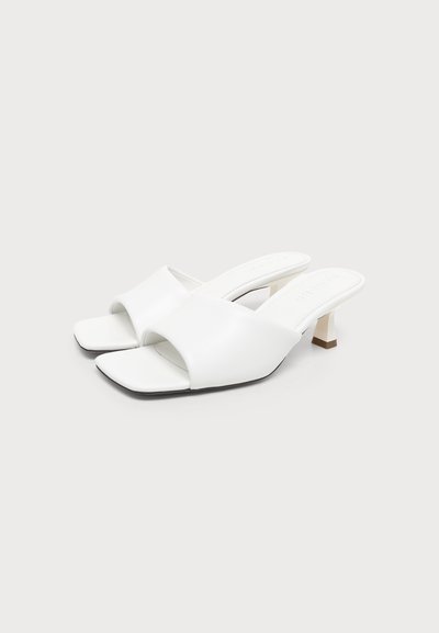 Sandales mule blanches à bout ouvert avec petits talons blocs et larges brides, présentées sur un fond clair uni.