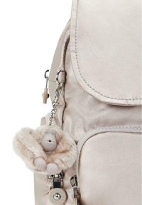 Jasnoróżowy nylonowy plecak z klapą; posiada puszysty breloczek w kształcie małpki i srebrne okucia; gładka tekstura i kompaktowy design.