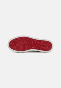 Semelle extérieure de sneaker blanche avec une semelle en caoutchouc rouge texturé, présentant des motifs circulaires et un logo, mettant en valeur une bordure blanche lisse.