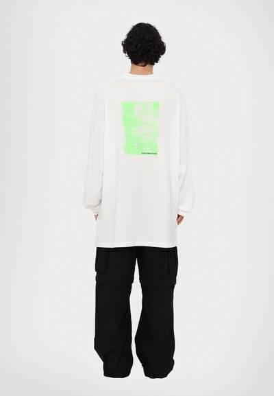 Camisa blanca de manga larga con un diseño gráfico verde en la parte posterior. Combinada con pantalones negros de corte holgado. Tela suave, ajuste relajado.