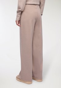 Kobieta w beżowych szerokich spodniach i dopasowanym swetrze, stojąca z jedną ręką swobodnie u boku na białym tle.