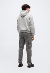 Νεαρός άνδρας που στέκεται με την πλάτη, φοράει ανοιχτό γκρι φούτερ με κουκούλα, σκούρα γκρι παντελόνια cargo και μαύρα αθλητικά παπούτσια σε απλό φόντο.