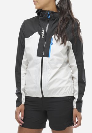 Femme portant une veste Millet noire et blanche avec des détails en zip bleu et un short noir, se tenant devant un fond uni.