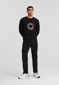 Felpa nera con logo rotondo e testo "KARL LAGERFELD". Abbinata a pantaloni neri e sneakers bianche. Texture liscia.