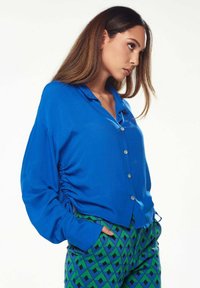 Blue button-up shirt with long sleeves, featuring gathered sides and a collar. Paired with patterned green and blue pants.