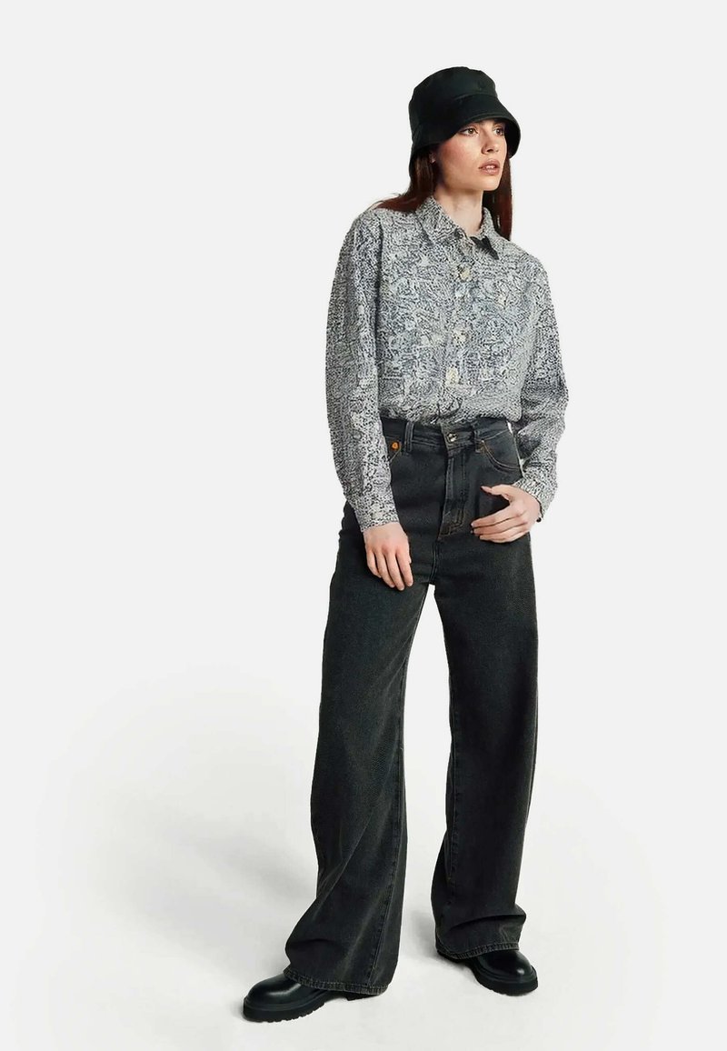 Camicia con bottoni dal motivo paisley grigio e bianco, abbinata a jeans neri a vita alta e gamba larga e stivaletti neri alla caviglia, con un cappello a secchiello nero.