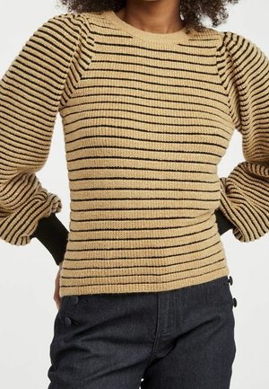 Žena nosící béžový a černý svetr s horizontálními pruhy, nabíranými rukávy a černými manžetami, v kombinaci s tmavými kalhotami s vysokým pasem.