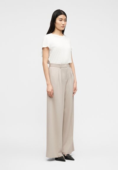 Γυναίκα που στέκεται φορώντας λευκό τοπ με κοντά μανίκια, μπεζ παντελόνι με ψηλή μέση και φαρδιά μπατζάκια, και μαύρα loafers με μυτερή μύτη, μπροστά σε λευκό φόντο.