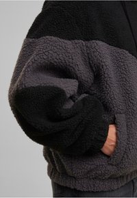 Inimese käsi kahe tooni musta ja tumeda halli tekstuuriga fliisjakki küljepocketis, millel on elastsed ääred ja varrukad.
