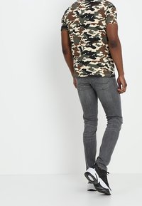 Koszulka moro w zielonym, brązowym i beżowym kolorze z krótkimi rękawami; zestawiona z dopasowanymi szarymi dżinsami i czarnymi trampkami z białymi akcentami.