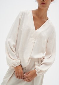 Helle beige Bluse mit tiefem V-Ausschnitt, langen Puffärmel mit elastischen Bündchen und einem weichen, strukturierten Stoff. Kombiniert mit passenden Hosen.