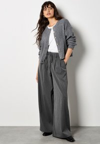 Cardigan grigio a coste con bottoni dorati, abbinato a pantaloni larghi grigi a righe. Camicia bianca semplice sotto. Scarpa nera completa il look.