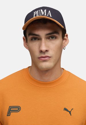 Pomarańczowy, fakturowany sweatshirt z okrągłym dekoltem, zestawiony z granatową czapką z białym napisem "PUMA" i kontrastującym wykończeniem.