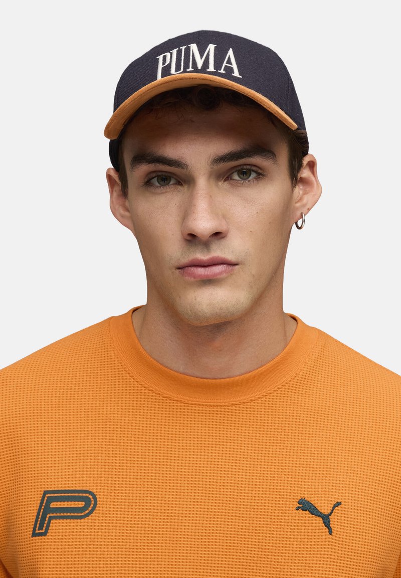Oranges Sweatshirt mit Struktur und Rundhalsausschnitt, kombiniert mit einer dunkelblauen Mütze, die das Wort "PUMA" in Weiß und einen kontrastierenden Unterseite zeigt.