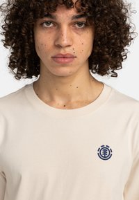 Beige Langarm-T-Shirt aus Baumwolle mit einem marineblauen Logodesign auf der Brust, rundem Halsausschnitt und glatter Textur.