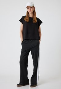 Zwarte cropped gebreide top met korte mouwen, gecombineerd met zwarte wijde broek met witte zijsstripes. Model draagt een witte pet en platte schoenen.