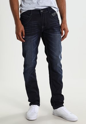 Hombre con jeans azul oscuro desgastados de corte ajustado, combinados con zapatillas blancas y una camiseta gris clara.