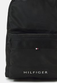 Zaino nero realizzato in nylon resistente con una tasca frontale con zip, caratterizzato da una striscia nera testurizzata e un piccolo logo rosso-bianco-blu.