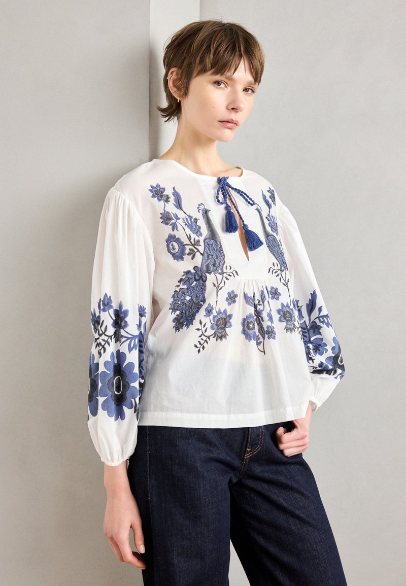 Weiße Bluse mit blauer Blumenstickerei, langen puffartigen Ärmeln und einem gebundenen Ausschnitt mit Quasten. Kombiniert mit dunklen Jeanshosen.