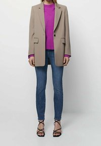 Beige Blazer, lila Pullover, blaue Jeans, schwarze Riemchen-Heels. Der Blazer hat Eingrifftaschen, einen Reverskragen und einen taillierten Schnitt.