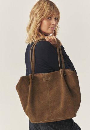 Braune Wildleder-Shopper-Tasche mit zwei stabilen Griffen. Verfügt über ein einfaches Design, oben etwas breiter, mit einem auffälligen Etikett auf der Vorderseite.