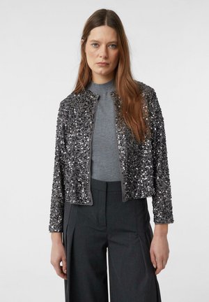 Woman with long brown hair wearing a gray sequin jacket over a gray top and dark gray wide-leg pants, standing against a plain background.
