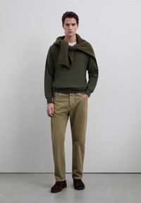 Conjunto masculino que incluye un suéter verde oscuro con cuello acanalado, pantalones caqui y botas de ante marrón. Diseño sencillo, paleta de colores neutros.