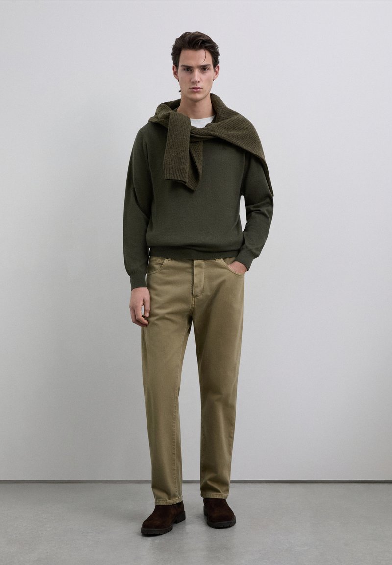 Conjunto masculino que incluye un suéter verde oscuro con cuello acanalado, pantalones caqui y botas de ante marrón. Diseño sencillo, paleta de colores neutros.